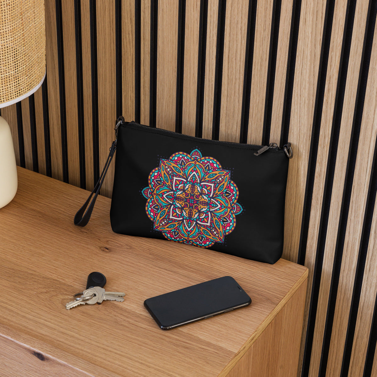 A black faux leather crossbody bag of Exclusivestan with a colorful mandala print, resting on a wooden table next to keys and a smartphone, with a slatted wood wall background.